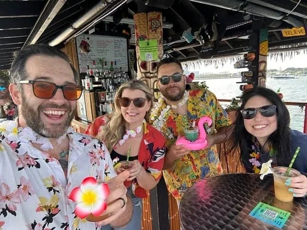 Group of friends wearing colorful shirts, flower leis, and drinking tropical cocktails on a Tiki Hut