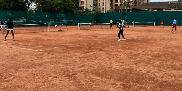 People playing tennis on a clay court with green fences and buildings in the background.