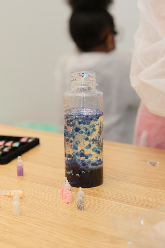 A clear bottle with blue glitter and small bottles of craft supplies on a table.