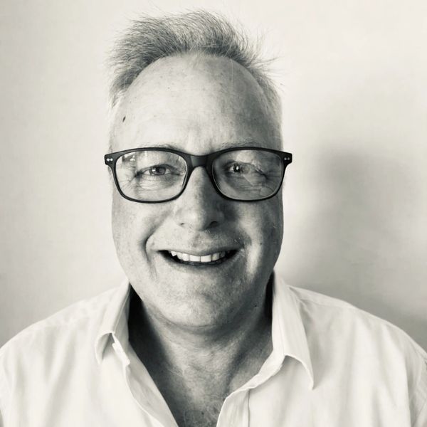 Smiling man with glasses and a white shirt against a plain background.