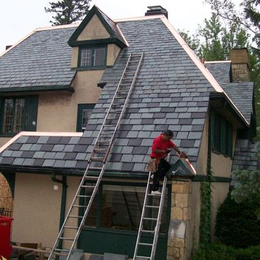 Worker installing or repairing roof shingles on a steep house roof using ladders.