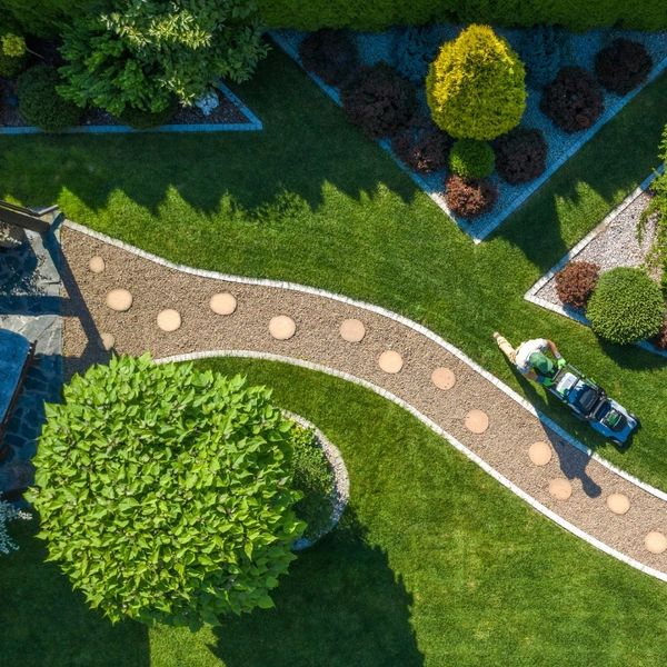 A person mowing a beautifully landscaped garden with trees and a winding path.