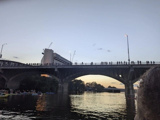 People stand on a bridge at dusk over a river with boats below.