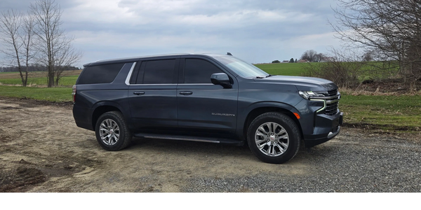 Dark gray Chevrolet Suburban parked on a gravel and dirt road with a green field background.