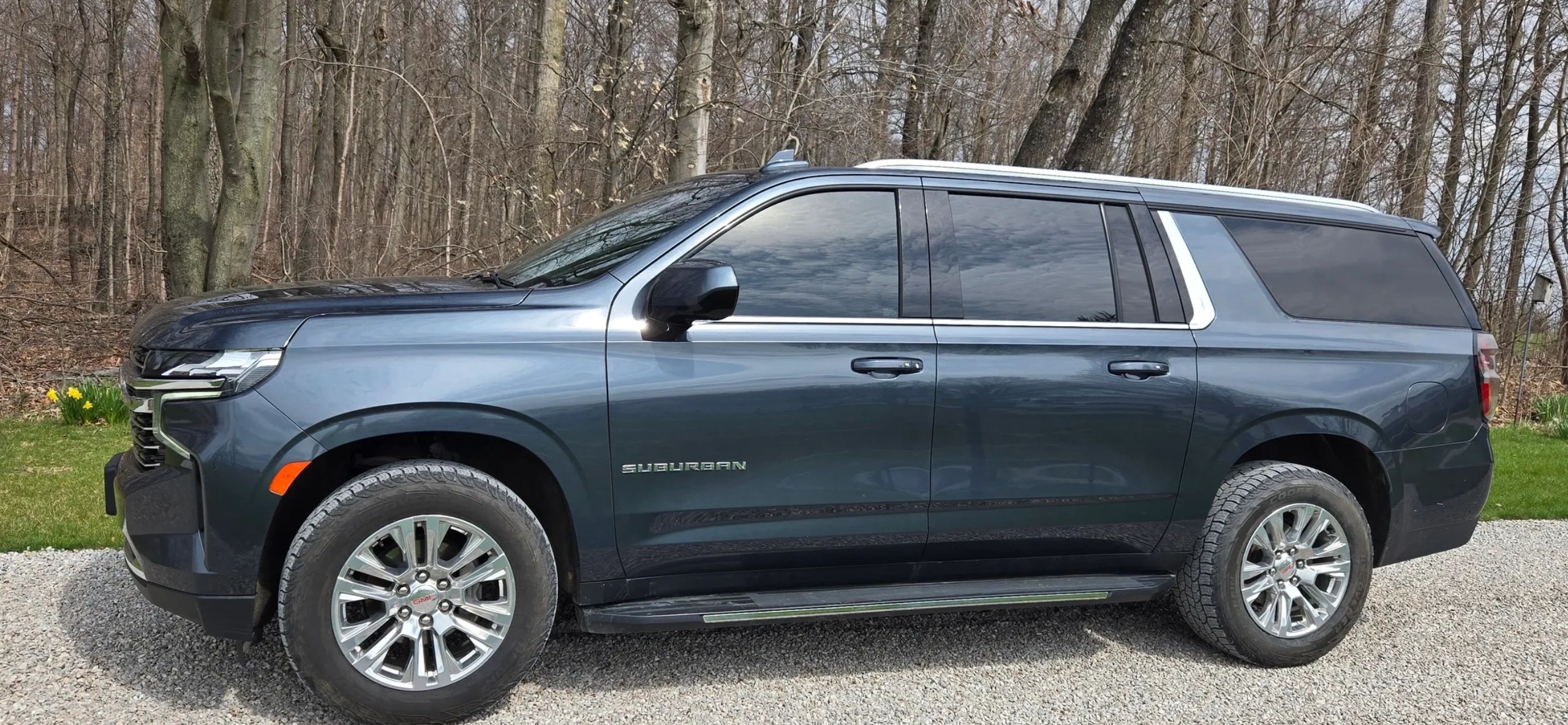 Dark blue GMC Suburban parked on gravel near a wooded area.