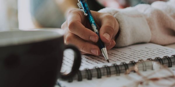 Writing using a pen and a notebook with a coffee mug on the side.