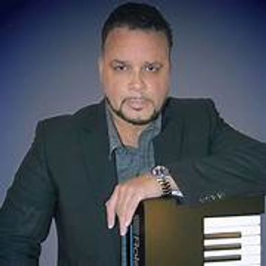 Man posing with a keyboard instrument, dressed in a dark blazer.