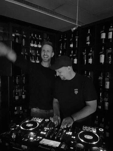 Zwei DJs beim Auflegen in einem dunklen, weinregal-umgebenen Raum.
