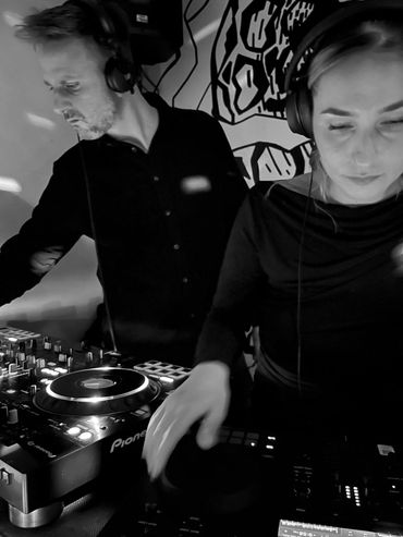 Zwei DJs bei der Arbeit, beide mit Kopfhörern und Bedienung von DJ-Equipment.