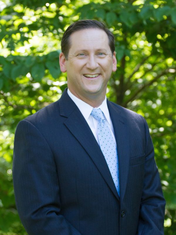 Man in a suit smiling outdoors with green foliage background.