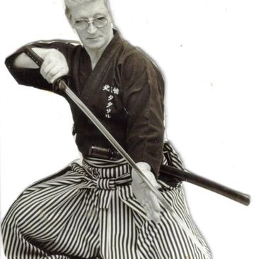 An elderly man dressed in traditional martial arts attire holding a katana.