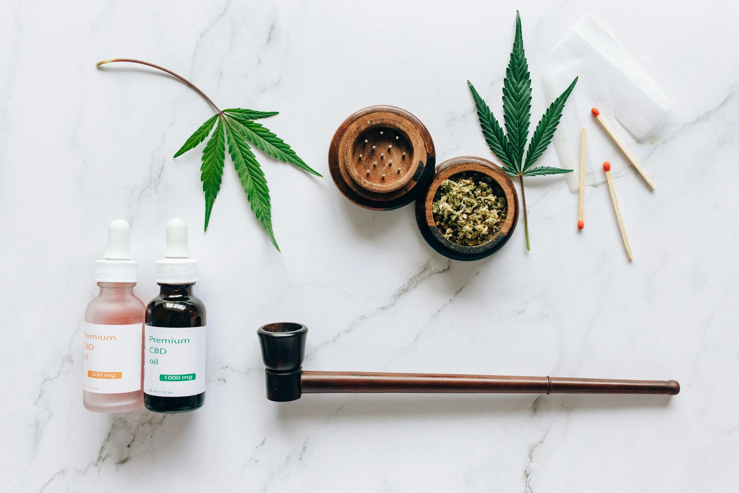 CBD oil bottles, cannabis leaves, grinder, pipe, and matches on a marble surface.