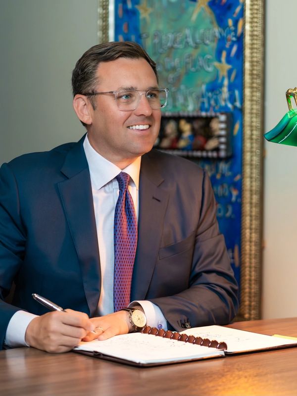 Man in suit writing at desk with green lamp and art in background.