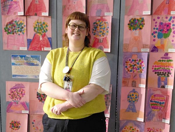 Person in yellow vest stands smiling in front of colorful children's artwork on a wall.