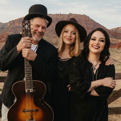Three people posing outdoors, one holding a guitar, all wearing black clothing and hats.