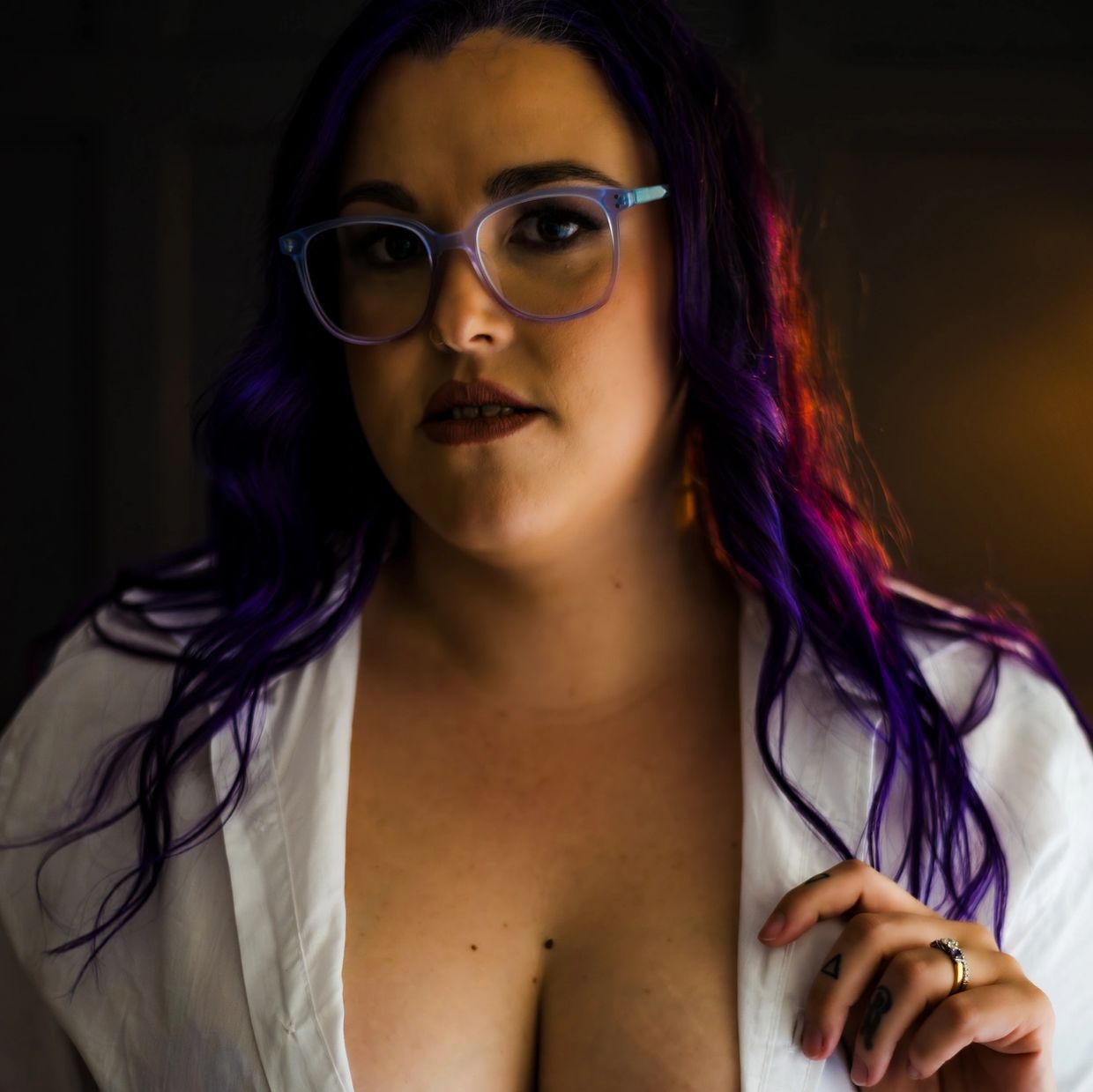 Woman with purple hair and glasses wearing a white top in warm lighting.
