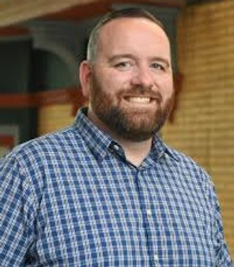 Smiling man with a beard wearing a blue plaid shirt indoors.