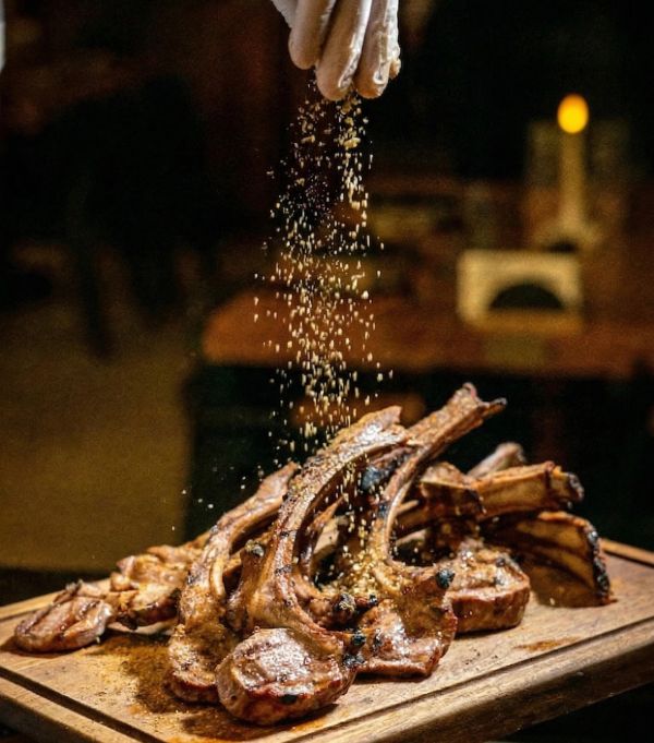 Hand sprinkling seasoning over grilled lamb chops on a wooden board.
