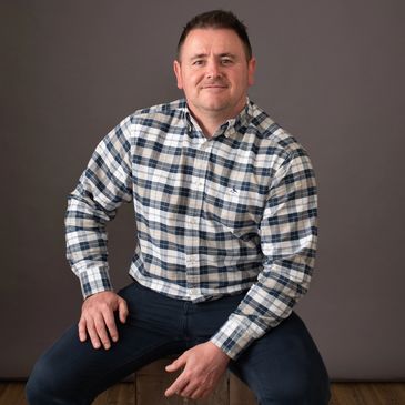 Man in a plaid shirt sitting on a wooden box against a gray backdrop.