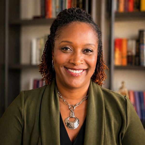 Smiling woman wearing a green jacket and silver necklace in a cozy room with bookshelves.