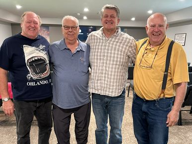 Four older men standing together indoors, smiling warmly.