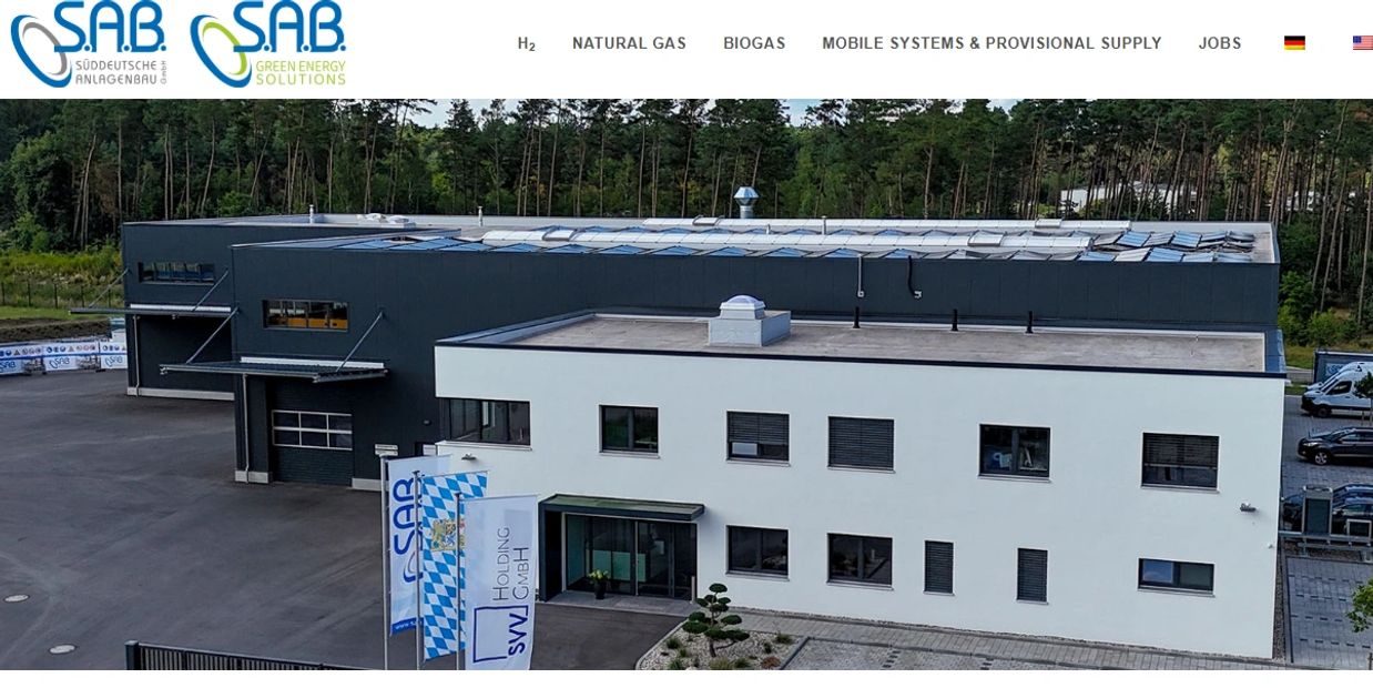 Modern industrial building surrounded by trees with company flags in front.