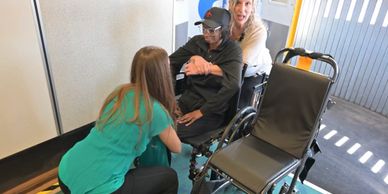 Three people assisting a woman in a wheelchair inside a building near an empty wheelchair.