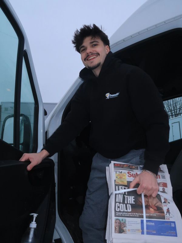 Smiling man holding newspapers stands by an open vehicle door.