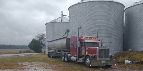 A red semi-truck parked near large grain silos on a cloudy day.