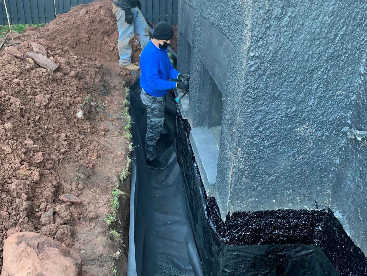 Two workers waterproofing a house foundation trench outdoors.