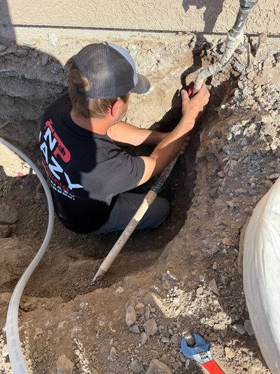 A worker repairs a pipe in a dirt trench beside a building.