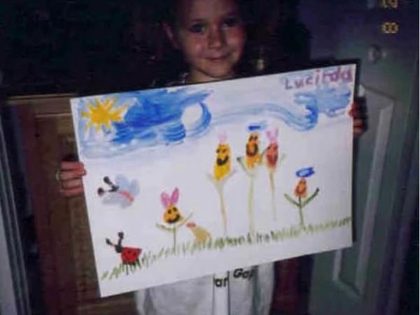 Child proudly displays a colorful drawing of flowers and insects under a bright sun.