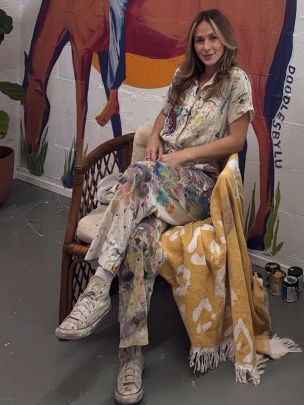 Woman in paint-splattered clothes sitting on a chair with a colorful backdrop.
