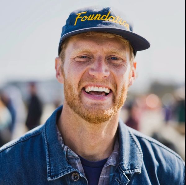 Smiling man with a beard wearing a blue cap and denim jacket.