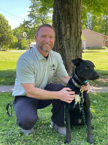 Man kneeling on grass holding a black dog on a leash in a sunny park.