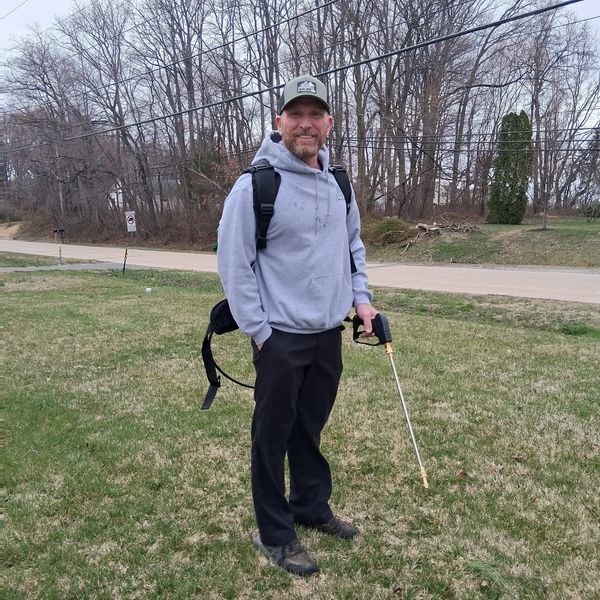 Man in gray hoodie holding a metal detector in a grassy area.