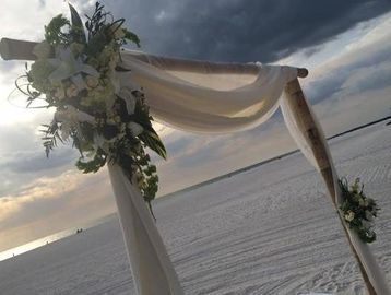 A rustic wedding arch decorated with flowers and fabric on a beach at sunset.
