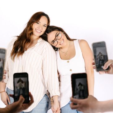 Two women smiling, holding hands, and being photographed by four smartphones.