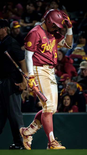 A baseball player in a red Trojans uniform adjusts his helmet during a game.