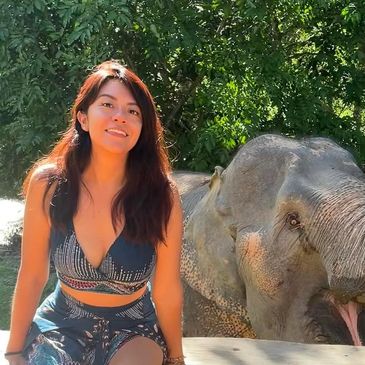 A woman smiling next to an elephant in a sunny outdoor setting.