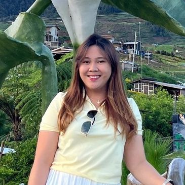 A smiling woman in a yellow shirt posing outdoors with greenery and hills in the background.