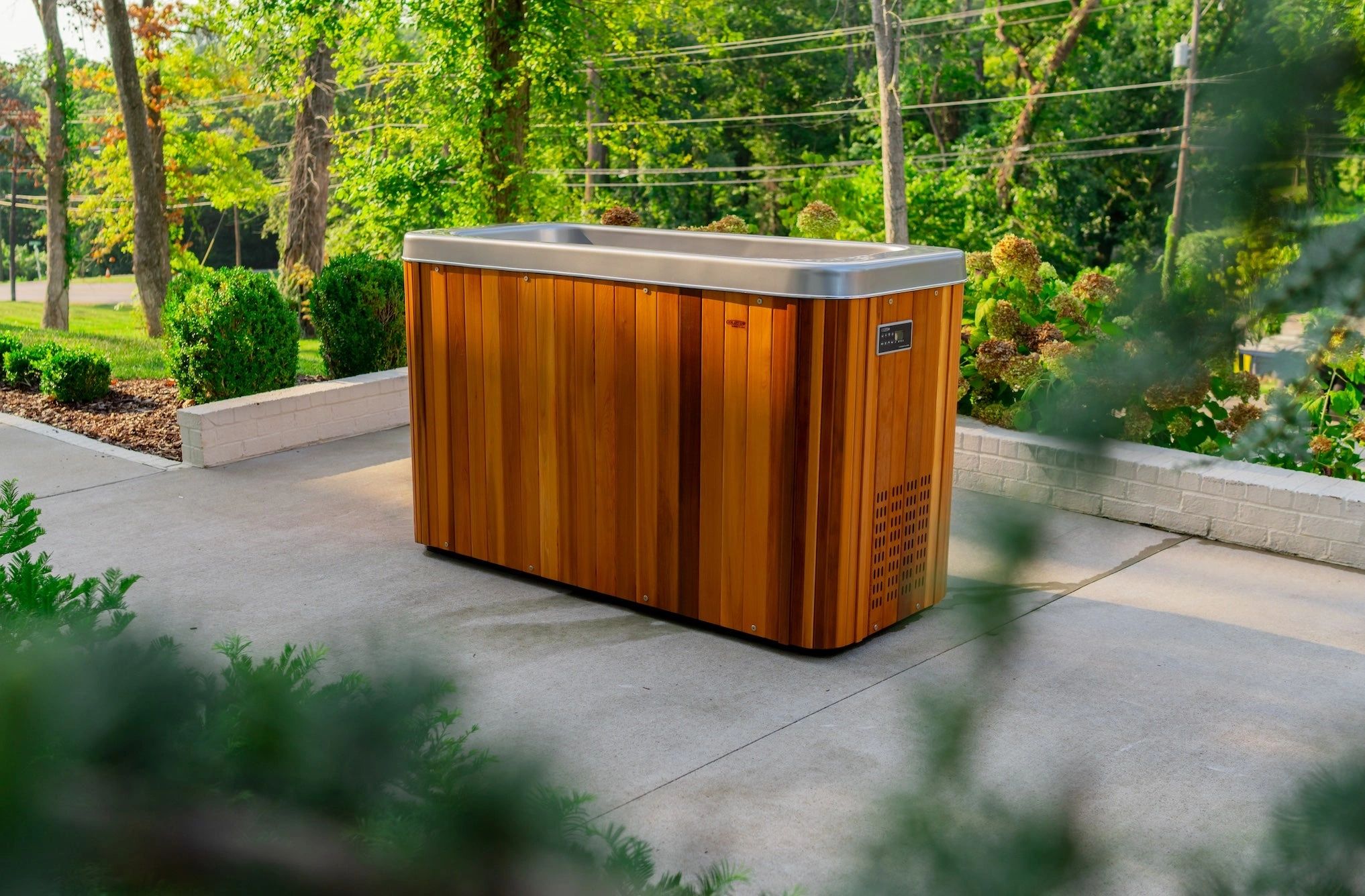 A wooden hot tub sits outdoors on a concrete patio surrounded by greenery.