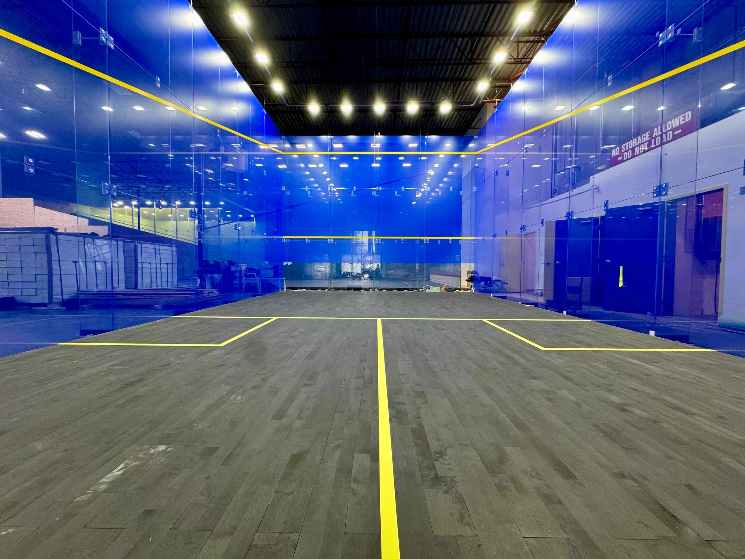 Indoor squash court with blue glass walls and wooden floor.