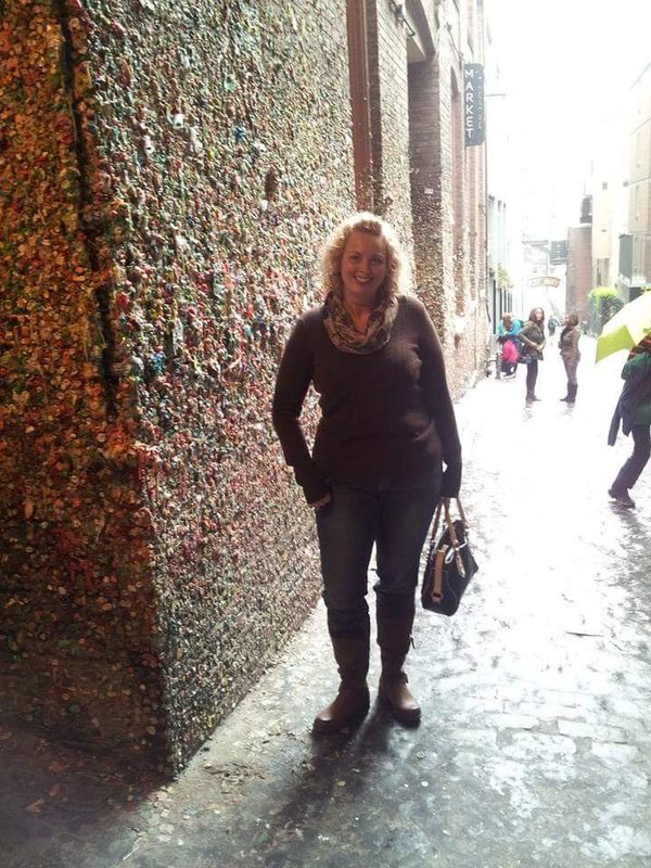 Woman stands near a wall covered in chewing gum in an alley.