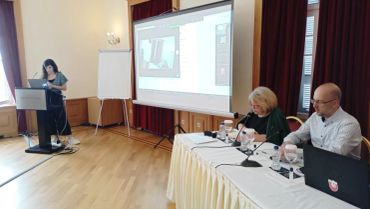 A speaker presenting to a panel and audience in a conference room.