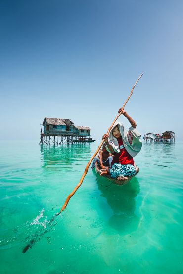 Child paddling a wooden boat near stilt houses on clear turquoise water.