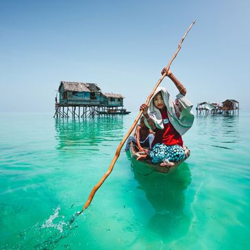 Person rowing a boat in clear turquoise water near stilt houses.