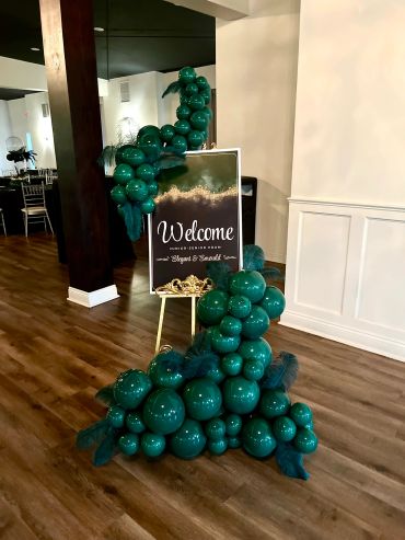 Welcome sign decorated with green balloons and foliage at an event venue entrance.