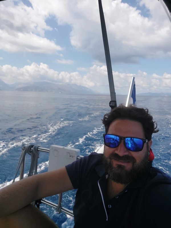 Man with sunglasses smiling on a boat with sea and mountains in the background.