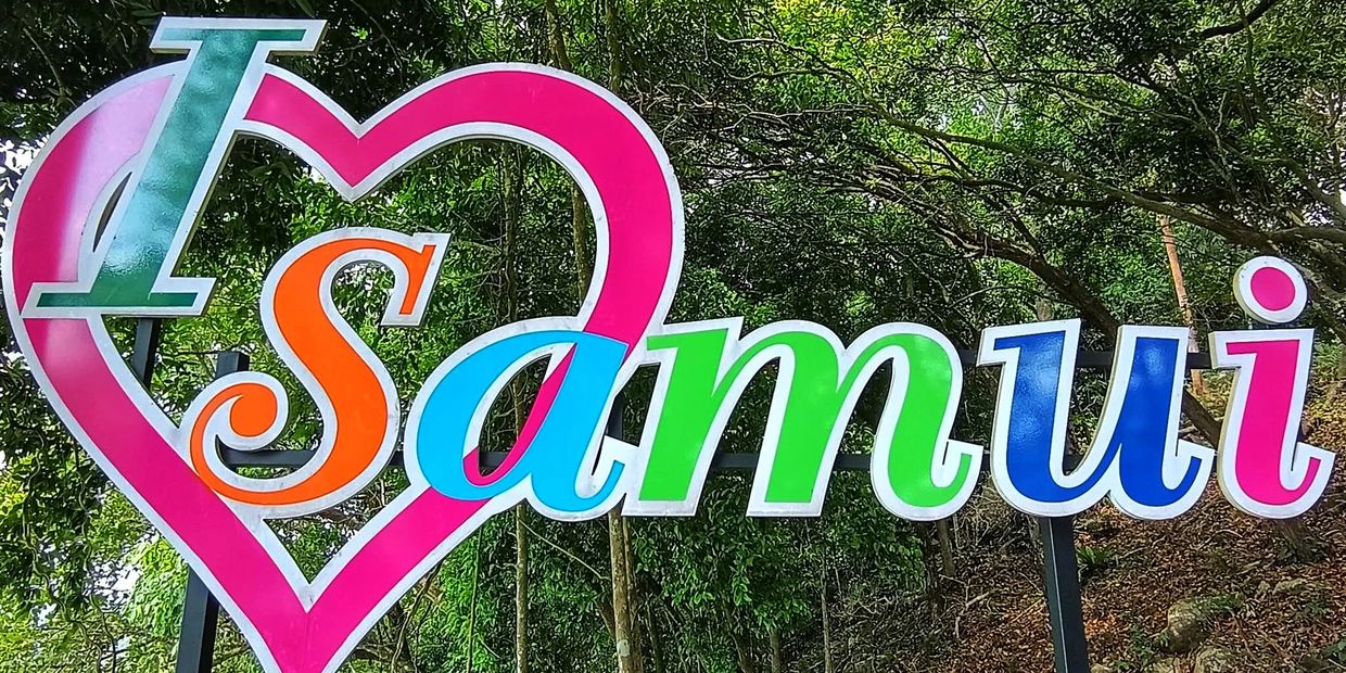 Colorful sign saying 'I love Samui' against a green forest background.